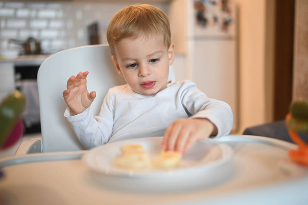 Little happy cute baby toddler boy blonde sitting on baby chair playing with banana. Baby facial expressions indoors at home kitchen interior with food. Healthy eating happy family childhood concept. - Foto, afbeelding
