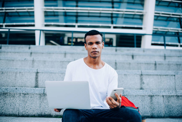Pensive African American male freelancer in casual outfit sitting on stairs on street and using laptop and smartphone in daytime - Fotó, kép