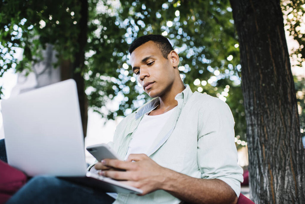 Low angle of confident young black man in casual outfit sitting on bag chair and working remotely on netbook while messaging on smartphone in street near trees in summer day - Fotó, kép
