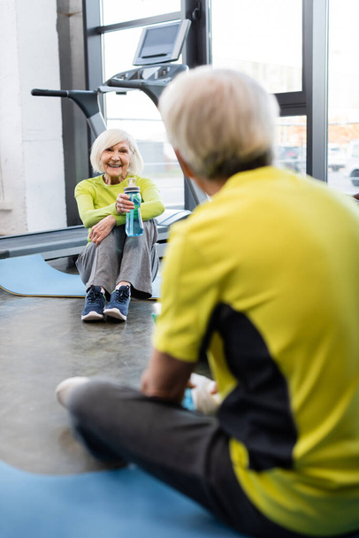 Uśmiechnięta starsza kobieta trzyma butelkę sportową w pobliżu niewyraźnego mężczyzny na siłowni  - Zdjęcie, obraz