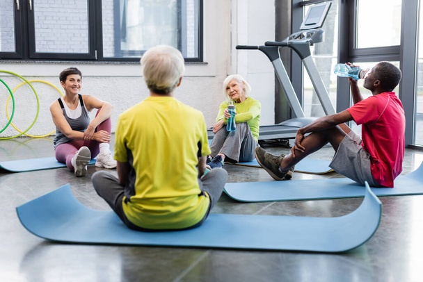 Positive multiethnic senior people with sports bottles resting on fitness mats in gym  - Photo, Image