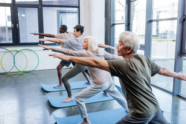 Seniorin in Kriegerpose praktiziert Yoga in der Nähe multiethnischer Menschen in Turnhalle   - Foto, Bild