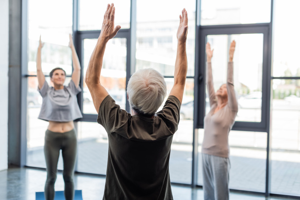 Senior man raising hands while practicing yoga near blurred friends in sports center  - Photo, Image