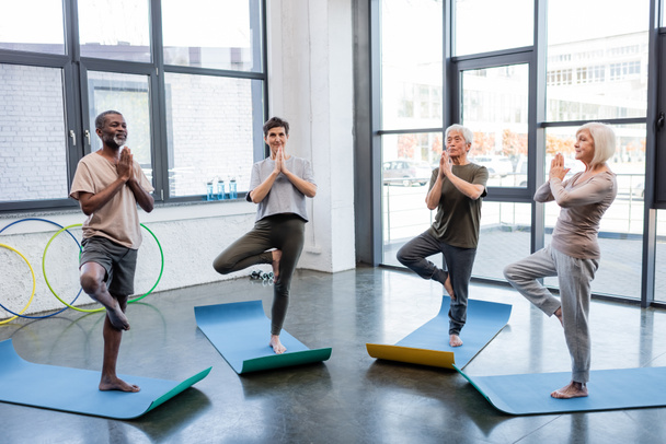 Interracial senior people standing in tree pose on yoga mats in sports center  - Photo, Image