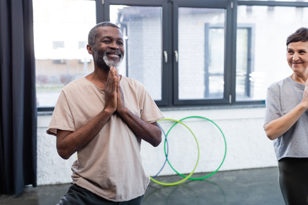 Sonriente hombre afroamericano practicando yoga cerca de un amigo mayor en un centro deportivo  - Foto, Imagen