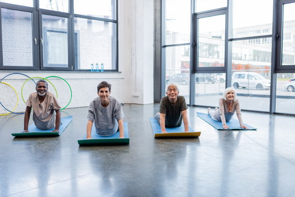 Smiling multicultural senior people looking at camera on yoga mats in sports center  - Foto, imagen