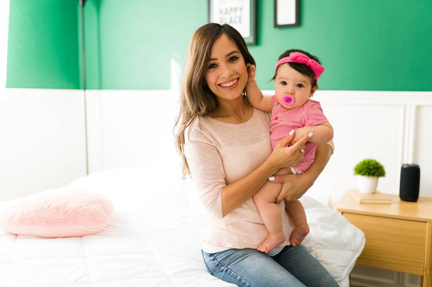 Attractive young mother carrying her four month baby girl in her arms while sitting in their bedroom - Photo, image