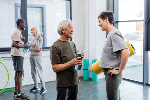 Smiling interracial people with sports bottle and fitness mat talking in gym  - Photo, Image