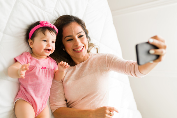 Attraktive Mutter macht ein Selfie mit ihrem fröhlichen süßen Baby-Mädchen, während sie es sich im Bett gemütlich macht - Foto, Bild