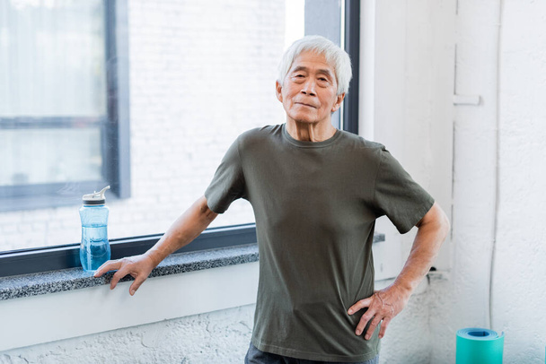 Grey haired asian man looking at camera near sports bottle in gym  - Photo, Image