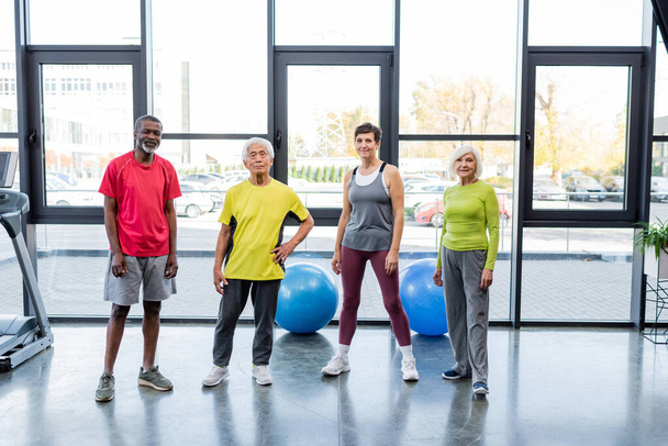 Positieve multi-etnische ouderen kijken naar camera in de buurt van fitnessballen en loopband in de sportschool  - Foto, afbeelding