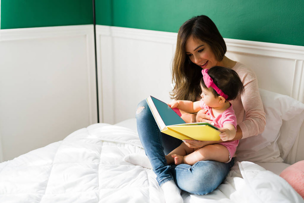 Lovely young mom reading a children's book to her cute baby girl while relaxing in bed together - Foto, immagini