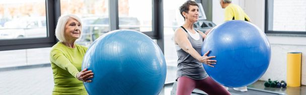 Grey haired sportswoman holding fitness ball in sports center, banner  - Фото, изображение