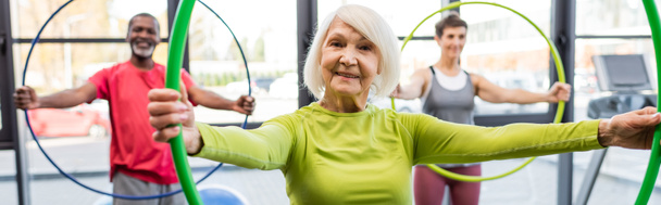 Lächelnde grauhaarige Frau mit Hula-Hoop-Reifen beim Training im Fitnessstudio, Banner  - Foto, Bild