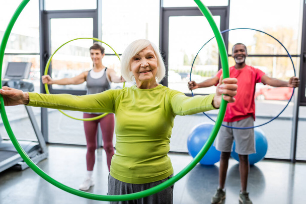 Starsza kobieta uśmiechnięta do kamery podczas treningu z hula hoop na siłowni  - Zdjęcie, obraz