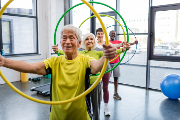Positive interrassische Menschen mit Hula-Hoop-Reifen und Blick in die Kamera im Fitnessstudio  - Foto, Bild