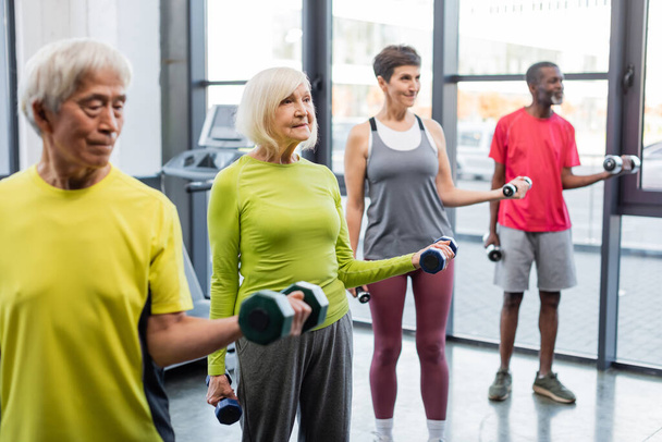 Smiling grey haired woman training with dumbbells near multicultural friends in gym  - Photo, Image