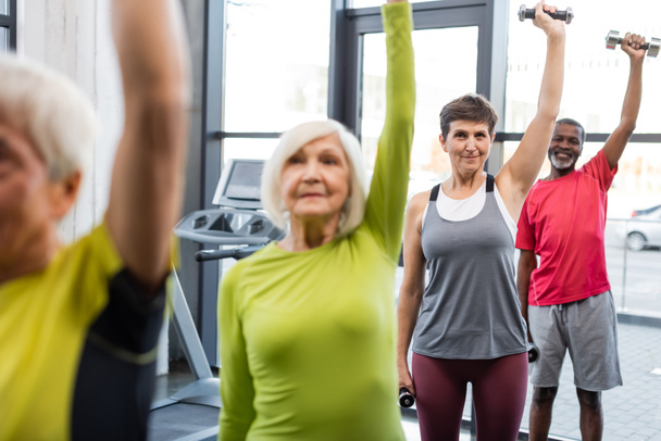 Mujer mayor haciendo ejercicio con pesas cerca de amigos multiétnicos en el gimnasio  - Foto, Imagen
