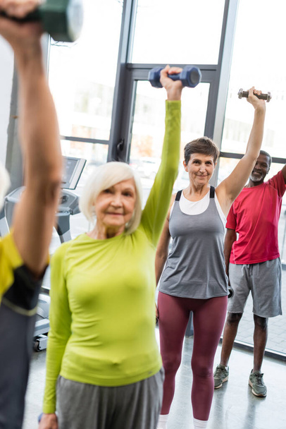 Interracial senior people looking at camera while training with dumbbells in sports center  - Photo, Image