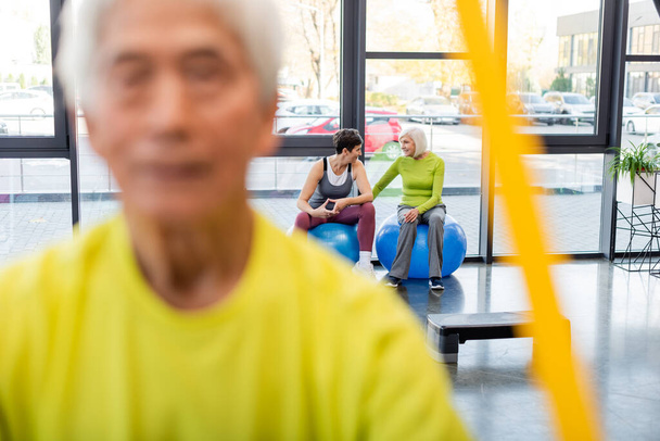 Uśmiechnięte starsze panie sportowe rozmawiające na piłkach fitness w pobliżu niewyraźnego treningu mężczyzn na siłowni  - Zdjęcie, obraz