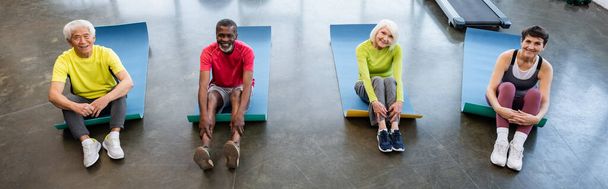 Vista de ángulo alto de la gente mayor interracial sonriente mirando la cámara en las esteras de fitness en el gimnasio, pancarta  - Foto, imagen