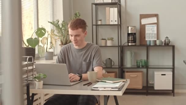 Moyen long de jeune homme caucasien aux cheveux roux assis au bureau le jour, travaillant sur un ordinateur portable et buvant du café dans une tasse - Séquence, vidéo