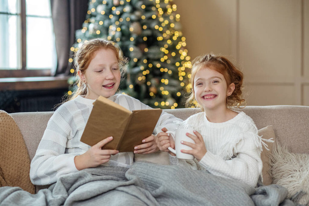 Two sisters are sitting on the couch and reading a book. Delicious hot cocoa. Children smile. Christmas Holidays. Warm white knitted sweaters. - Foto, Imagem