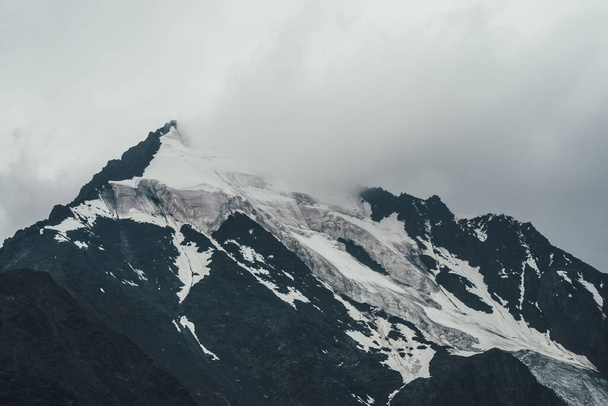 Μινιμαλιστικό μονόχρωμο ατμοσφαιρικό ορεινό τοπίο με μεγάλη χιονισμένη κορυφή βουνού σε χαμηλά σύννεφα. Φοβερό minimal σκηνικό με παγετώνα σε βράχους. Μπλε λευκό ψηλό βουνό πυραμίδα με χιόνι στα σύννεφα. - Φωτογραφία, εικόνα