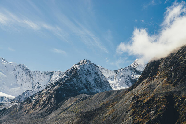 Ατμοσφαιρικό αλπικό τοπίο με υψηλό χιονισμένο βουνό με κορυφώθηκε κορυφή κάτω από τα σύννεφα cirrus στον ουρανό. Μεγάλο χιόνι σκεπασμένο με ήλιο. Χαμηλό σύννεφο σε μαύρα βράχια και λευκό χιόνι μυτερή κορυφή στο φως του ήλιου - Φωτογραφία, εικόνα