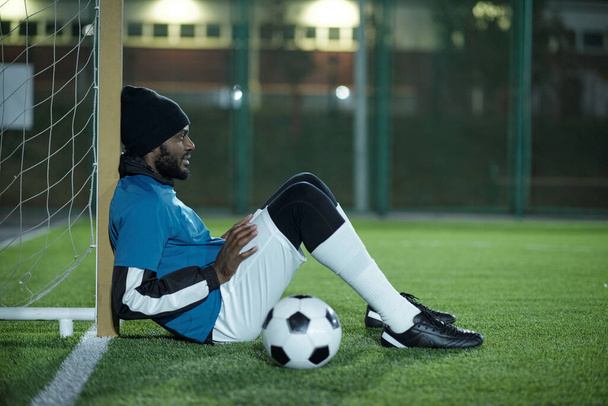 Young African footballer sitting by net on green lawn - Photo, Image