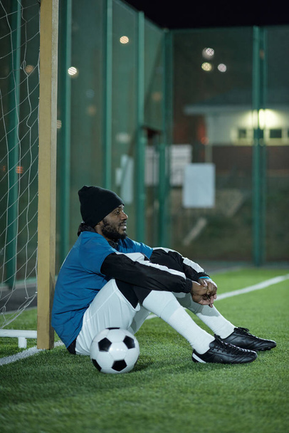 Νεαρός ξεκούραστος ποδοσφαιριστής κάθεται στο δίχτυ μετά από σκληρή προπόνηση - Φωτογραφία, εικόνα