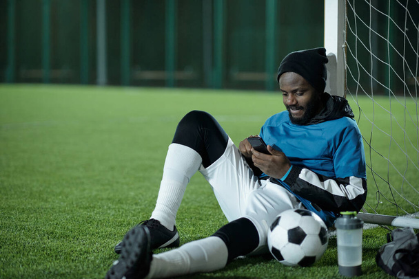 Young restful sportsman sitting by net and scrolling in smartphone - Zdjęcie, obraz
