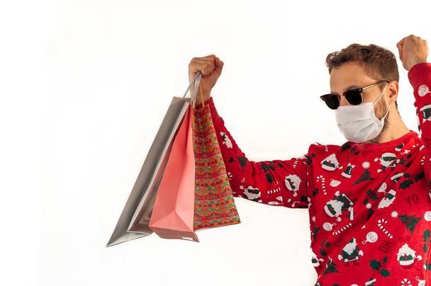 Młody biały człowiek w czerwonym świątecznym swetrze, okularach przeciwsłonecznych i masce trzymający torby z rękami w górze, odizolowany na białym tle i przestrzeni do kopiowania. Koncepcja zakupów. - Zdjęcie, obraz