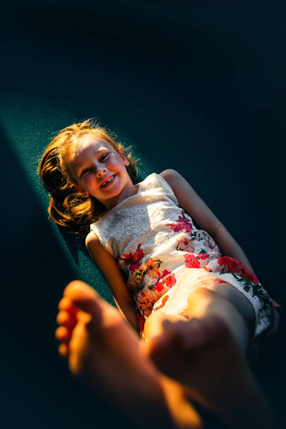 A little girl lies on the floor in a beam of light and stretches her leg towards the camera. - Fotó, kép