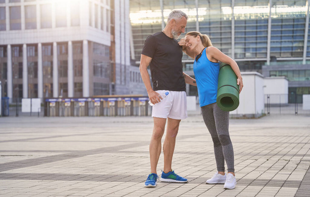 Belo casal de meia idade em sportswear de pé ao ar livre com tapete de ioga após o treino matinal na cidade - Foto, Imagem