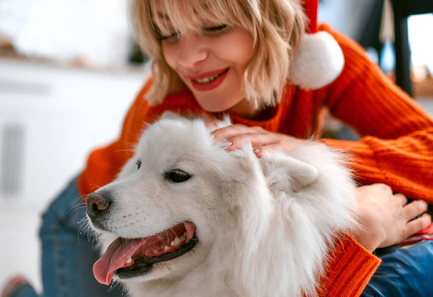 Merry Christmas and Happy New Year! Cute woman in santa hat hugging her pet and friend fluffy white dog. - Фото, изображение