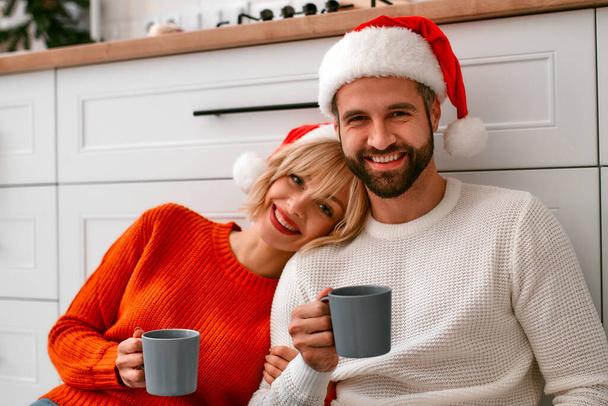 Καλά Χριστούγεννα και Καλή Χρονιά! Νεαρό ζευγάρι με κέρατα ταράνδου στην κουζίνα πίνουν καφέ ενώ κάθονται στο πάτωμα εν αναμονή των διακοπών. - Φωτογραφία, εικόνα