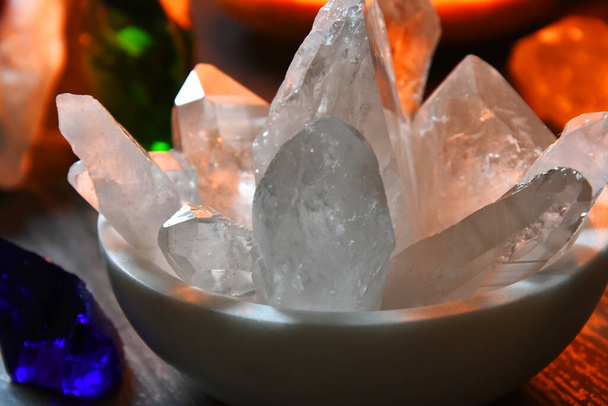 Una imagen de un tazón blanco lleno de una variedad de cristales de cuarzo claro con una lámpara de sal rosa brillante en el fondo.  - Foto, imagen