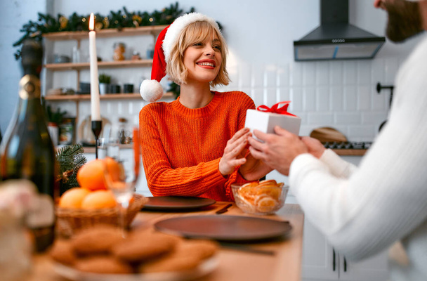 Καλά Χριστούγεννα και Καλή Χρονιά! Ένα νεαρό ερωτευμένο ζευγάρι με καπέλα Σάντα στην κουζίνα σε ένα ρομαντικό εορταστικό δείπνο δίνουν ο ένας στον άλλο δώρα, περιμένοντας τις διακοπές. - Φωτογραφία, εικόνα