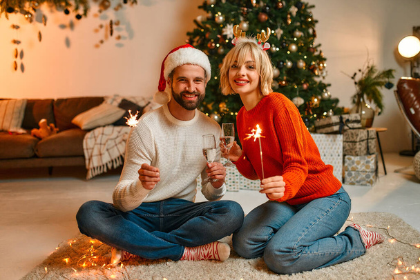Hyvää joulua ja onnellista uutta vuotta! Rakastava pari joulupukin hatuissa ja poronsarvissa istuu lattialla joulukuusen lähellä tähtisamppanjan ja lasien kanssa odottamassa lomaa.. - Valokuva, kuva