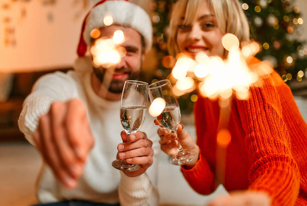メリークリスマスとハッピーニューイヤー!サンタ・ハットの素敵なカップルと、クリスマスツリーの近くの床に座っているトナカイの釣り人、シャンパンの輝きとガラス、休日を待っています. - 写真・画像