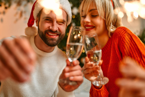 Καλά Χριστούγεννα και Καλή Χρονιά! Ένα ερωτευμένο ζευγάρι με καπέλα και κέρατα ταράνδων κάθεται στο πάτωμα κοντά σε ένα χριστουγεννιάτικο δέντρο με σπίθες και ποτήρια σαμπάνιας, περιμένοντας διακοπές. - Φωτογραφία, εικόνα