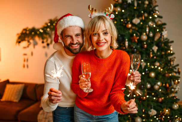 Joyeux Noël et bonne année ! Un couple aimant dans des chapeaux de Père Noël et des bois de renne assis sur le sol près d'un arbre de Noël avec des étincelles et des verres de champagne, attendant des vacances. - Photo, image