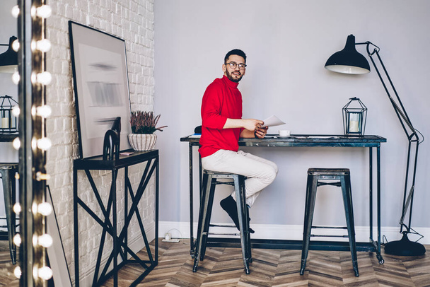 Caucasian man with paper documents sitting on chair in modern apartment with stylish interior and looking at camera, hipster guy in optical eyeglasses holding bills at home posing during weekend - Fotó, kép