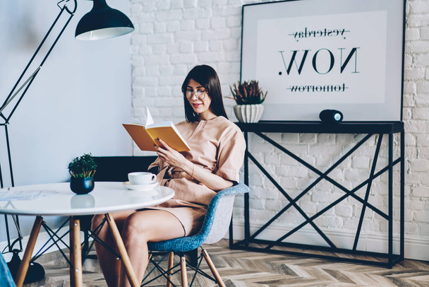Caucasian hipster girl in stylish eyewear for vision correction reading interesting literature book during leisure weekend in modern apartment,young woman interested in book plot while resting in home - Foto, Imagen