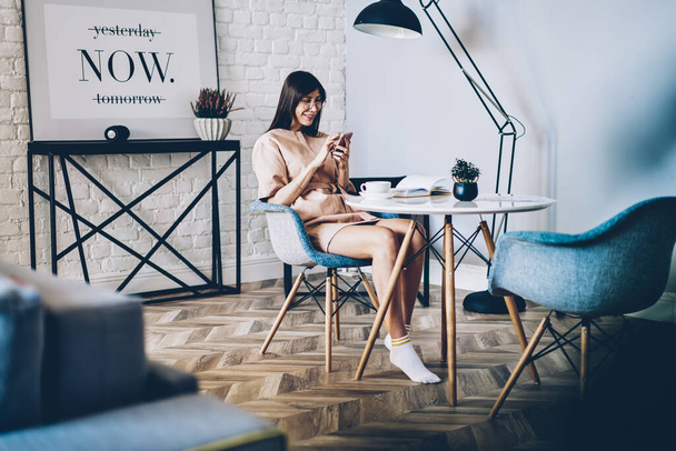 Pretty female blogger enjoying mobile messaging during leisure time in home apartment with good wifi internet, millennial woman in eyeglasses browsing content publication via application on cellular - Foto, Imagem