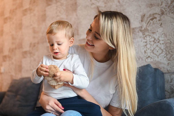 Little cute baby toddler boy blonde sitting on mothers arms. Beautiful young mom and son playing spend time together indoors at home with toys. Healthy happy family childhood concept. - Foto, Imagen