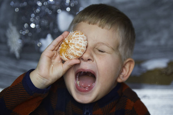 Porträt eines niedlichen, fröhlichen Kleinkindes. Junge spielt mit Mandarinen zwischen Weihnachtsdekoration. Festlich-stimmungsvolles Zuhause für Silvester. Ästhetisches gemütliches Zuhause. Fröhlicher kleiner Junge. Fröhliches Konzept.  - Foto, Bild