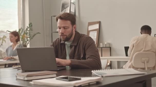 Середній план молодого бородатого кавказького чоловіка-менеджера сидячи за столом в офісі, використовуючи ноутбук, а потім дивлячись і посміхаючись на камеру - Кадри, відео