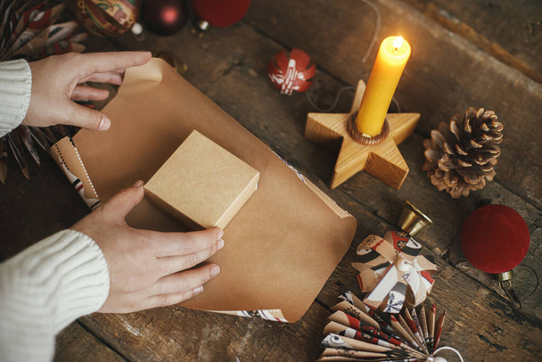 Pakuję świąteczny prezent. Ręce owijania pudełko na prezent w świątecznym papierze rzemieślniczym na rustykalnym drewnianym stole ze świecą, nożyczkami i ozdobami. Atmosferyczny obraz. Przygotowanie ferii zimowych - Zdjęcie, obraz